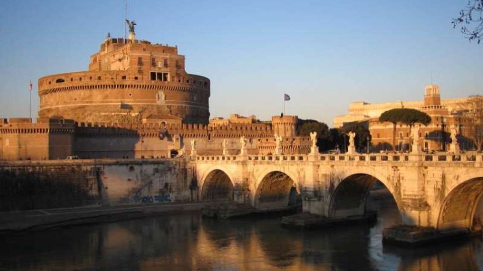 Veduta di Castel Sant’Angelo, fortezza cilindrica in muratura, con il Ponte Sant’Angelo in primo piano caratterizzato da archi in pietra e statue lungo la balaustra. Il ponte attraversa il fiume Tevere, con riflessi sull’acqua e cielo sereno sullo sfondo.