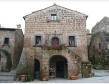 Facciata di una casa in pietra in un borgo storico, con grande arco d’ingresso al piano terra, finestre simmetriche e balconcino con porta ad arco e vasi di fiori. Pavimentazione in pietra e edifici adiacenti in muratura su entrambi i lati.