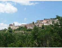 Vista panoramica di un borgo collinare con case in pietra e tetti in laterizio, disposto in salita lungo il pendio. In primo piano si vede vegetazione verde e alberi, con cielo azzurro parzialmente nuvoloso sullo sfondo.
