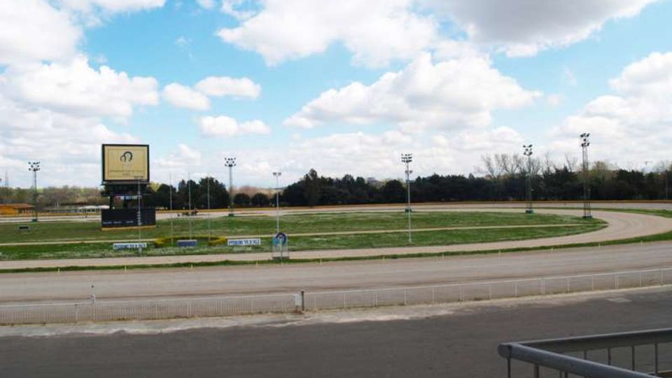 Veduta di un ippodromo all’aperto con pista ovale in terra battuta, area centrale erbosa e recinzioni lungo il bordo. Sullo sfondo si vedono un grande tabellone elettronico e diversi pali con fari, con alberi all’orizzonte sotto un cielo parzialmente nuvoloso.