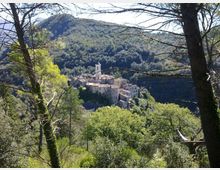 Veduta da un pendio boscoso su un piccolo borgo in pietra arroccato su una collina, con un campanile bianco che spicca tra i tetti. Sullo sfondo si estendono montagne coperte di foreste, incorniciate da alberi e rami in primo piano.