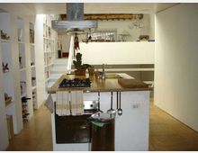 Cucina moderna in ambiente open space con isola centrale in legno dotata di piano cottura, lavello e cappa in metallo sospesa. Ai lati si vedono scaffalature bianche a giorno e una parete divisoria in vetro satinato, con pavimento in legno chiaro e arredi minimalisti.