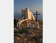 Antica torre costiera intonacata di bianco su un promontorio roccioso affacciato sul mare, raggiunta da una rampa e una scalinata con parapetti. In primo piano si vede una grande croce nera su piedistallo e alcuni muretti e lampioni lungo il percorso.