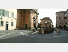 Piazza lastricata in pietra con una fontana circolare al centro e paletti dissuasori intorno. Sullo sfondo si vedono edifici storici dai toni chiari e la facciata laterale di una chiesa con alti pilastri e dettagli in pietra.