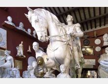 Interno di uno studio o magazzino d’arte con pareti rosse e soffitto a travi, pieno di statue e busti in gesso disposti su scaffali e a terra. Al centro domina una grande scultura equestre bianca, circondata da numerose altre sculture di varie dimensioni.
