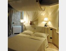 Camera da letto in stile rustico con letto matrimoniale, biancheria chiara e baldacchino in tessuto appeso al soffitto. Ai lati ci sono lampade da comodino su una testiera in muratura, con tende e parete decorata sullo sfondo.
