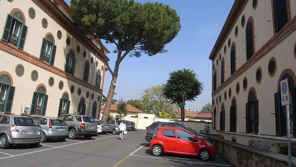 Cortile-parcheggio tra due edifici a più piani con facciate chiare, finestre ad arco e piccoli oblò circolari lungo le pareti. Al centro si vedono alberi alti e diverse auto parcheggiate, con strada asfaltata e segnaletica a terra.