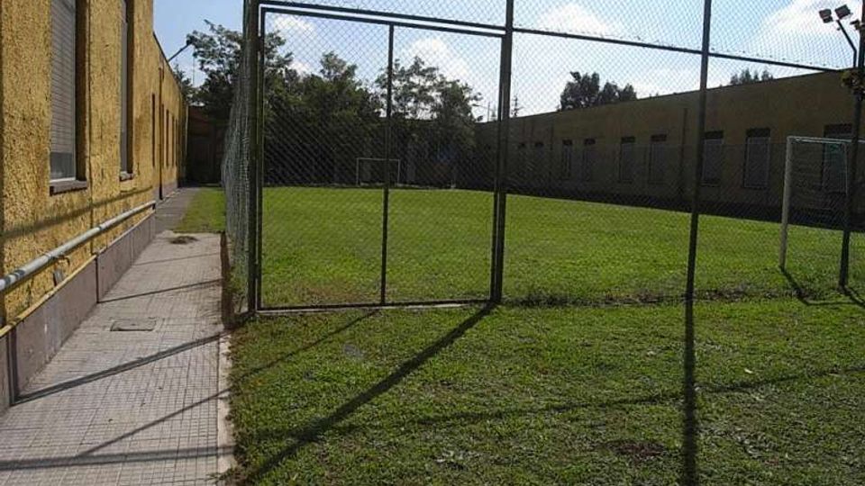 Campo da calcio in erba recintato con rete metallica, con porte bianche alle estremità. A sinistra corre un marciapiede lungo un edificio giallo e sullo sfondo si vedono altri edifici bassi e alberi sotto un cielo sereno.