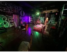 Interno di uno spazio scenico tipo teatro o studio con pavimento scuro lucido, illuminazione colorata verde e viola e pareti ricoperte da grandi murales in stile street art. Al centro c’è una pedana con corde sospese e cuscini, circondata da sedie vintage e da alcuni elementi d’arredo e luci da palco.