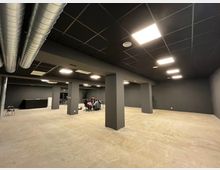 Ampio spazio interno tipo ufficio o sala polifunzionale con pareti e colonne grigio scuro, pavimento in cemento e soffitto nero a griglia con luci a pannello. Sono visibili condotti di aerazione metallici a vista e un lungo bancone sul lato sinistro.