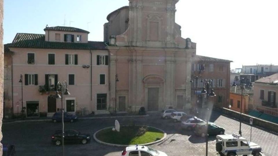 Veduta di una piazza urbana con piccola rotonda centrale erbosa e strada con auto parcheggiate, di fronte a una chiesa storica con facciata barocca in muratura chiara e portale centrale. Intorno si trovano edifici residenziali a più piani con finestre e tetti in tegole, illuminati da luce diurna.