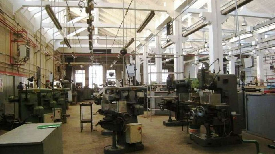 Interno di un’officina industriale con ampio capannone, soffitto alto a travi bianche e lucernari, illuminato da lampade sospese. Macchinari e attrezzature meccaniche sono disposti in file su pavimento in cemento, con grandi finestre sul fondo.
