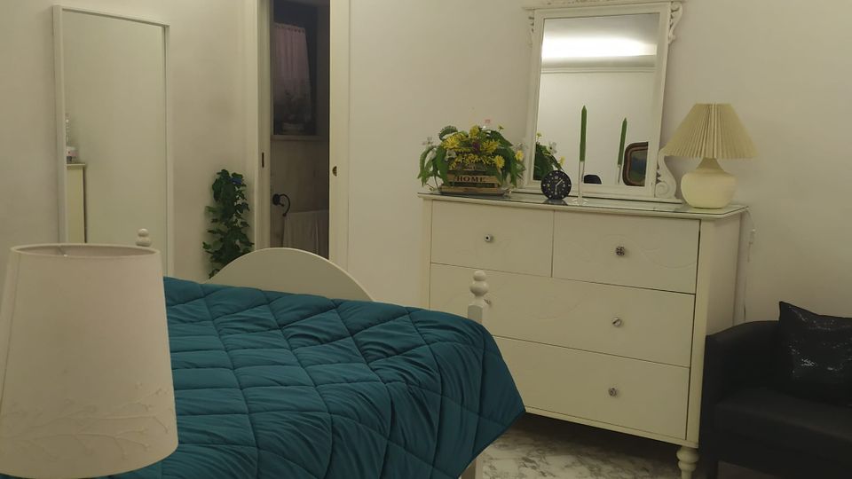 Camera da letto luminosa con pareti bianche, pavimento in marmo chiaro e letto con copriletto blu. Sono visibili una cassettiera bianca con grande specchio decorato, armadio, lampade da tavolo e una porta aperta verso un altro ambiente.