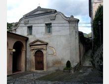 Facciata di una piccola chiesa in intonaco chiaro con timpano curvilineo, croce in sommità e portale in pietra con porta in legno. Davanti c’è un cortile lastricato e, sul lato destro, una stretta salita tra edifici che conduce verso l’alto.