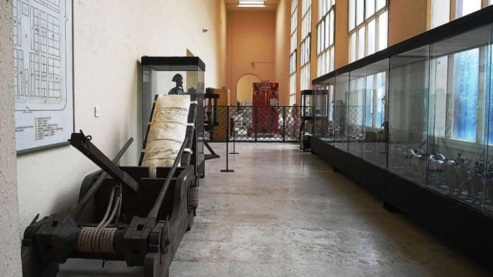 Corridoio museale lungo e stretto con alte finestre a griglia su un lato e grandi teche espositive in vetro. In primo piano si vede una grande macchina in legno e metallo tipo catapulta, con altri reperti e pannelli informativi lungo le pareti e una recinzione in fondo al passaggio.