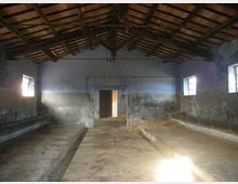 Interno di una stalla o fienile vuoto con tetto a capriate in legno a vista e pareti intonacate usurate. Il pavimento in cemento presenta due lunghi rialzi laterali e luce naturale entra da piccole finestre e da una porta centrale sul fondo.
