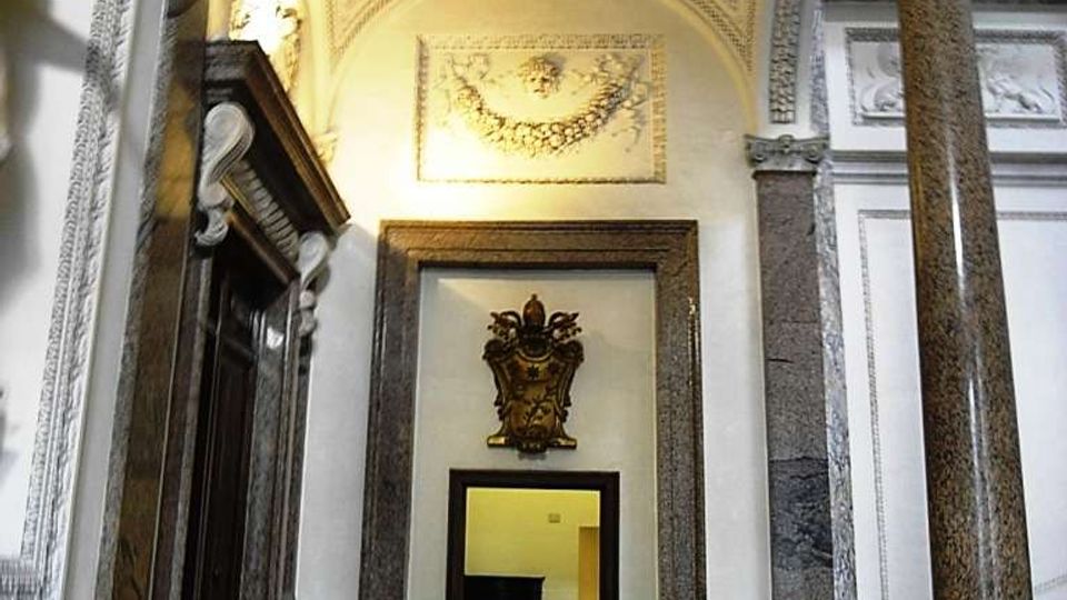 Interno di un edificio storico con atrio a volta decorata a cassettoni e stucchi, colonne in pietra e cornici in marmo attorno a una porta centrale. Pavimento in marmo con motivo geometrico e scalinate laterali sotto archi ornamentali.