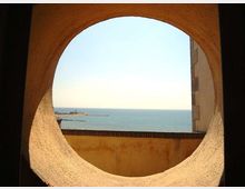 Veduta del mare attraverso un grande foro circolare in una parete in muratura, con bordi intonacati e un muretto in primo piano. All’orizzonte si vede una diga o molo con un piccolo faro e il cielo limpido sullo sfondo.