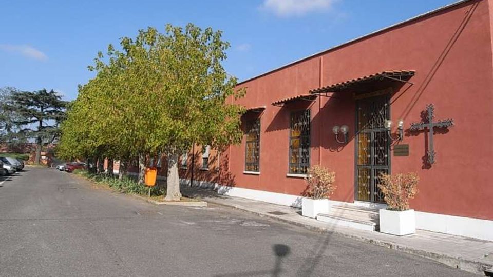 Edificio basso e allungato con facciata rosso mattone, finestre con inferriate e ingresso con piccolo portico, affacciato su una strada asfaltata. Davanti ci sono alberi lungo il marciapiede, alcune auto parcheggiate e un lampione, con cielo sereno sullo sfondo.