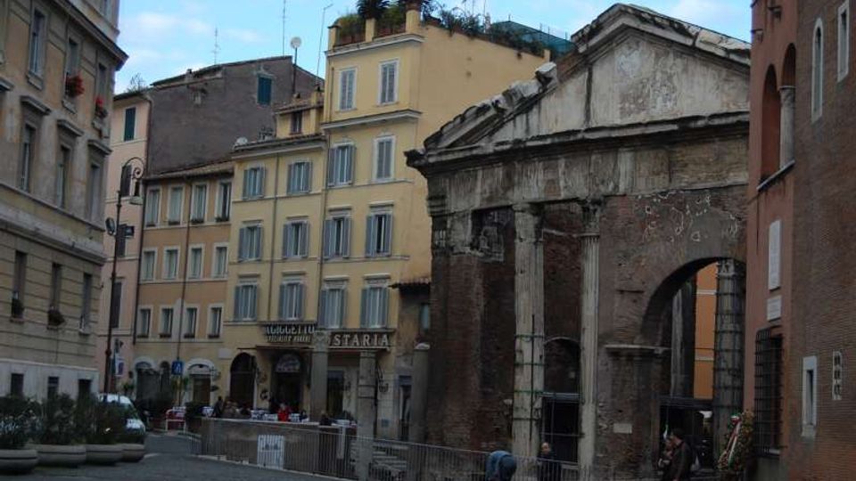 Piazza urbana con pavimentazione in pietra, circondata da palazzi storici dai toni giallo e beige con finestre regolari. In primo piano si vedono i resti di un’antica struttura in mattoni con colonne e frontone parzialmente crollato, delimitata da transenne.