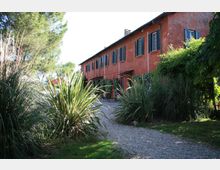 Edificio rurale a due piani con facciata intonacata color terracotta e persiane scure, affacciato su un vialetto in ghiaia. Intorno si trova un giardino con cespugli fitti, piante ornamentali e alberi alti che incorniciano l’ingresso.