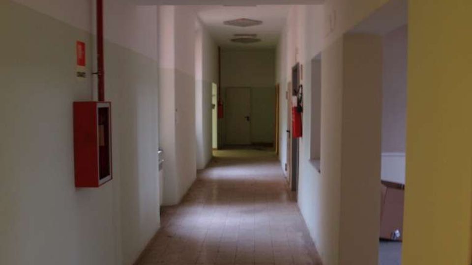 Corridoio interno lungo e stretto con pavimento in piastrelle, pareti bicolore e plafoniere a soffitto. Sulle pareti sono presenti cassette antincendio rosse ed estintori, con aperture laterali e una porta in fondo al passaggio.