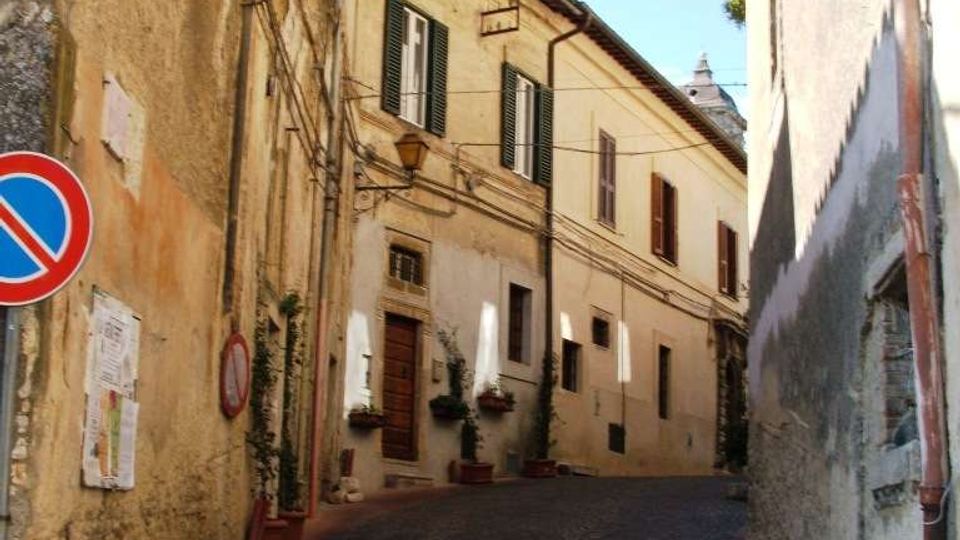 Strada stretta in salita con pavimentazione in ciottoli tra edifici storici in intonaco chiaro, con persiane, porte e vasi alle finestre. Sulla parete a sinistra sono visibili una lanterna a muro e un cartello di divieto di sosta, con cavi sospesi lungo la via.