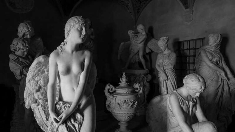 Interno di una galleria o deposito di sculture con statue in marmo disposte in un ambiente poco illuminato. In primo piano si vede una statua femminile con ali, mentre sullo sfondo compaiono altre figure classiche, un grande vaso decorato e archi con soffitto a volta.