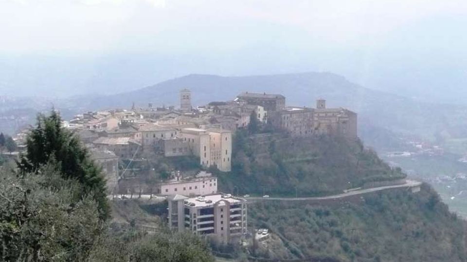 Veduta panoramica di un borgo storico su una collina, con edifici in pietra e una torre campanaria, affacciato su una valle nebbiosa. Una strada serpeggiante corre lungo il pendio tra vegetazione e campi sottostanti.