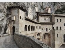 Complesso monastico in pietra addossato a una parete rocciosa, con portico ad archi, finestre strette e tetti in coppi. In primo piano un camminamento con parapetto conduce a un ingresso ad arco e a corpi di fabbrica con piccoli campanili e rosone circolare.