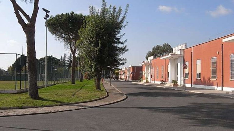 Strada asfaltata curva in un’area urbana tranquilla, con alberi alti e un’area verde recintata a sinistra. A destra si vedono edifici bassi color rosso mattone con ingressi bianchi e finestre allineate lungo il marciapiede.