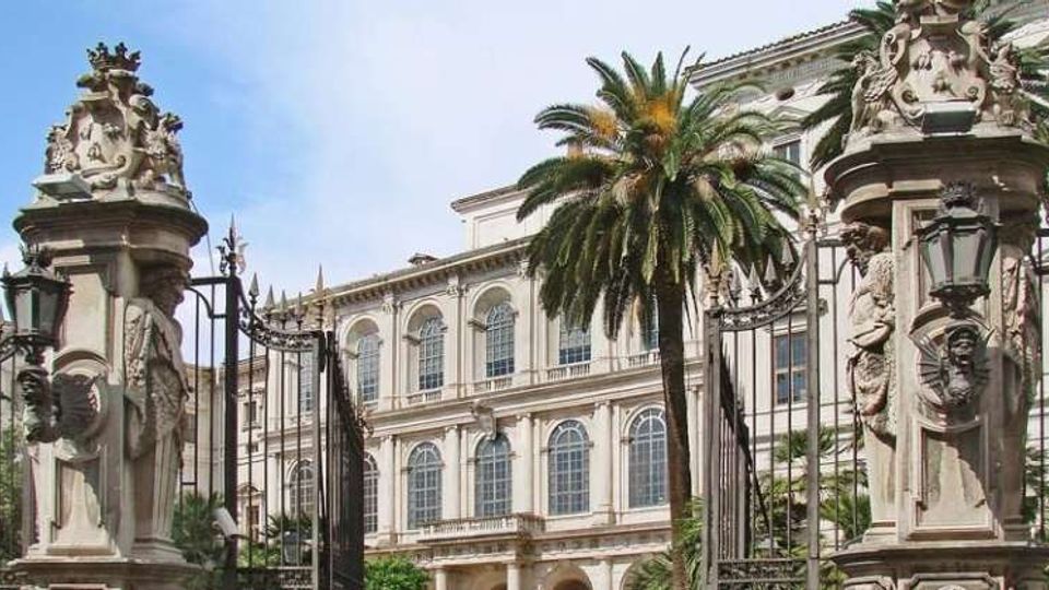 Ingresso monumentale con cancelli in ferro battuto tra due pilastri in pietra decorati con sculture e lanterne. Sullo sfondo si vede un grande edificio storico a più piani con finestre ad arco e palme nel cortile.