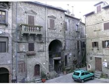 Facciate di edifici in pietra in un borgo storico, con finestre a persiane, balcone con balaustra e un grande arco passante al centro. Piccola piazzetta lastricata con scale laterali e un’auto parcheggiata vicino agli ingressi.