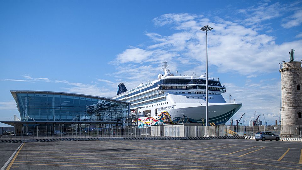 Terminal portuale moderno con facciata vetrata e tetto curvo, accanto a una grande nave da crociera ormeggiata. Sul lato destro si vede una torre in pietra tipo faro e, in primo piano, un ampio piazzale asfaltato con segnaletica.