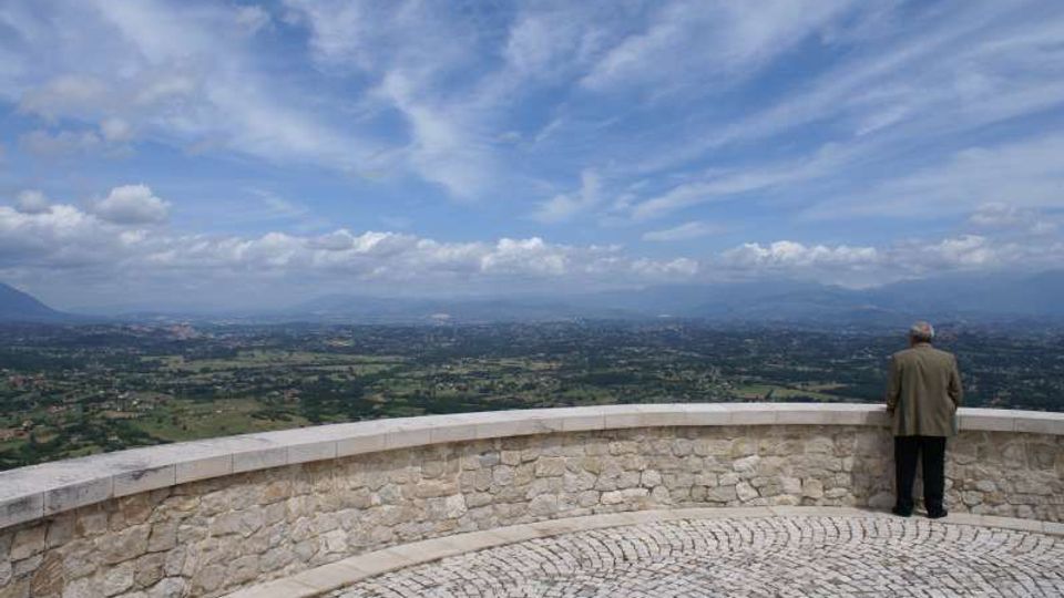 Terrazza panoramica in pietra con parapetto semicircolare e pavimentazione in ciottoli, affacciata su una vasta valle verde con campi e centri abitati. Sullo sfondo si vedono montagne lontane sotto un ampio cielo azzurro con nuvole sparse.