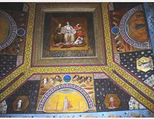 Soffitto decorato di un interno storico con riquadri dipinti e cornici dorate, con un grande pannello centrale figurativo e motivi ornamentali geometrici. Intorno si vedono medaglioni e pannelli con scene e figure su fondo blu e oro, tipici di una volta affrescata.
