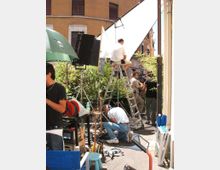 Set cinematografico all’aperto in una strada urbana, con grandi pannelli riflettenti e luci su cavalletti, scale a pioli e cavi elettrici sul pavimento. Sullo sfondo si vedono facciate di edifici e vegetazione lungo il bordo della strada.