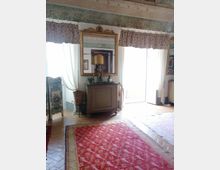 Salotto in stile classico con pavimento in parquet e grande tappeto rosso, credenza con specchio dorato e decorazioni, e soffitto con fregi ornamentali. Ampie porte-finestre con tende bianche e mantovane fiorate illuminano la stanza.
