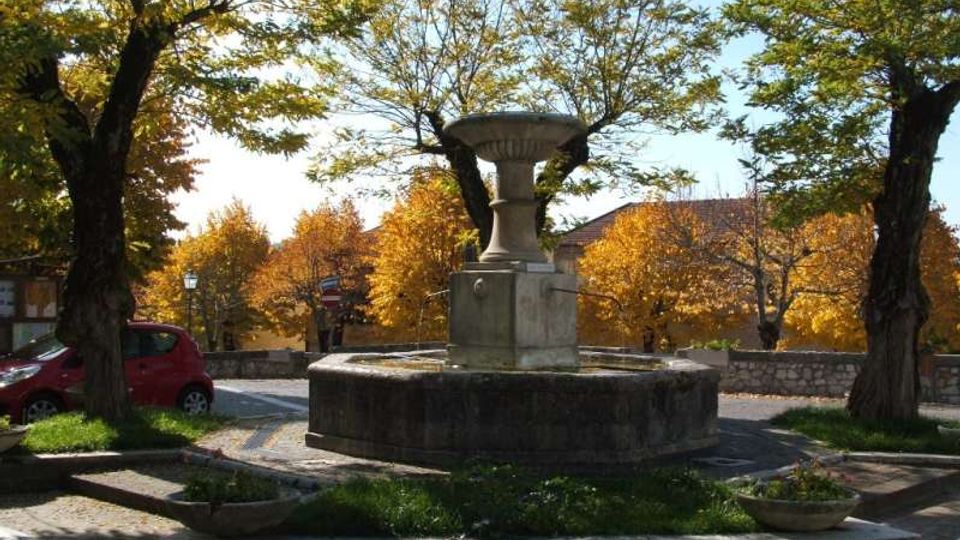 Fontana in pietra con vasca circolare al centro di una piccola piazza pavimentata, circondata da aiuole e alberi. Sullo sfondo si vedono un muretto in pietra e case basse tra vegetazione autunnale.