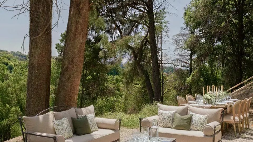 Area lounge all’aperto in un bosco, con divani imbottiti e tavolino in ferro battuto su ghiaia sotto alberi alti. Sullo sfondo si vede un lungo tavolo da pranzo apparecchiato con sedie in legno, circondato da vegetazione e colline.