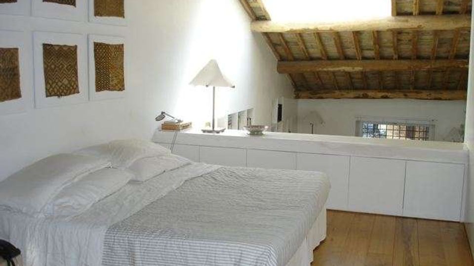 Camera da letto mansardata con pavimento in legno, letto matrimoniale con biancheria chiara e pareti bianche decorate con quadri. Il soffitto è in travi di legno a vista con lucernario, e lungo il lato c’è un mobile basso con lampada.
