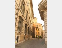 Strada stretta acciottolata in un centro storico, fiancheggiata da alti palazzi in pietra con finestre con inferriate e cornici modanate. Sullo sfondo si vedono edifici color ocra e un lampione, con una piccola piazzetta con siepi.