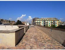 Camminamento sopraelevato con pavimentazione in mattoni e muretti laterali intonacati, che conduce verso edifici residenziali. Sullo sfondo si vedono palazzi a più piani e un cielo sereno con alcune nuvole.