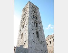 Torre campanaria medievale in pietra chiara vista dal basso, con più ordini di bifore e trifore ad arco e muratura massiccia. Sul lato destro si intravede parte di un edificio in pietra con finestre ad arco sotto un cielo azzurro.