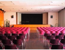 Sala auditorium con file di sedie rosse disposte a platea e corridoio centrale, rivolta verso un palco rialzato con pannellatura in legno e fondale scuro. Soffitto con illuminazione a incasso, pareti chiare e tende laterali.