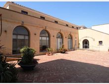 Cortile lastricato in mattoni con vasi di piante, affiancato da un edificio intonacato color ocra con grandi finestre ad arco. Sullo sfondo si vede un ingresso ad arco in un’ala adiacente e un cielo sereno sopra i tetti in coppi.