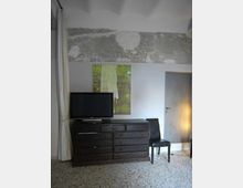 Interno di una stanza con pareti bianche e una fascia alta di intonaco grezzo a vista, pavimento in graniglia e tenda chiara sul lato sinistro. In primo piano ci sono una cassettiera scura con un televisore sopra, una sedia nera e una porta chiusa sulla destra.