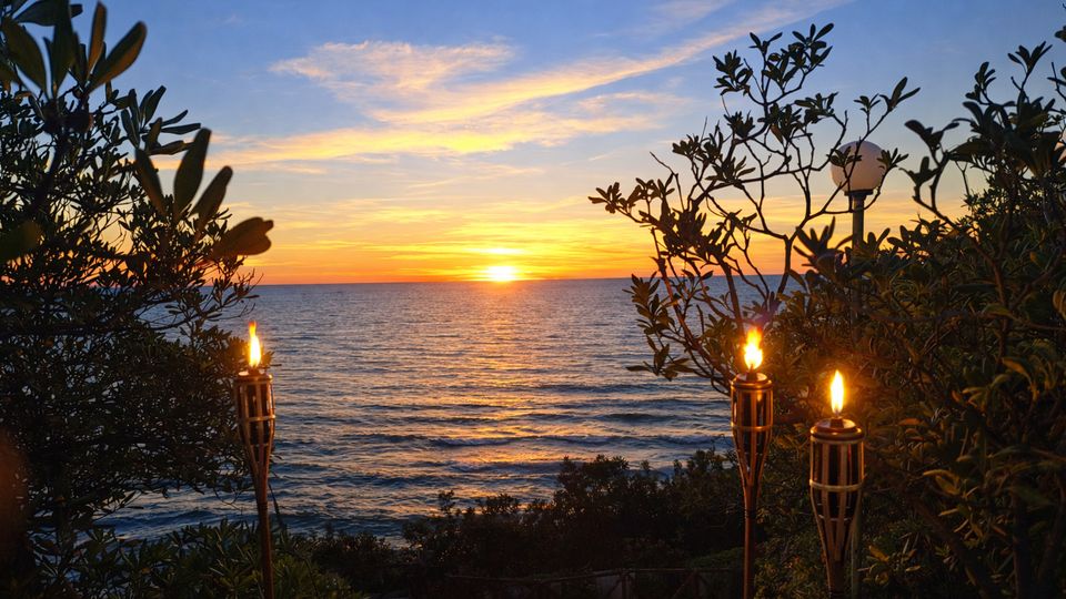 Vista sul mare al tramonto da un giardino costiero, con vegetazione in primo piano e torce accese su pali lungo il bordo. All’orizzonte il sole scende sull’acqua calma sotto un cielo con nuvole sottili.