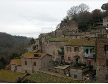 Borgo collinare con case in pietra addossate tra loro e tetti spioventi in coppi, disposte su più livelli lungo un pendio. Sullo sfondo si vedono alberi spogli e una valle boscosa sotto un cielo coperto.