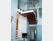 Interno moderno a doppia altezza con scala a sbalzo in legno e ringhiera in metallo che conduce a un soppalco. Pareti bianche, pavimenti in vetro e un camino incassato completano l’ambiente.